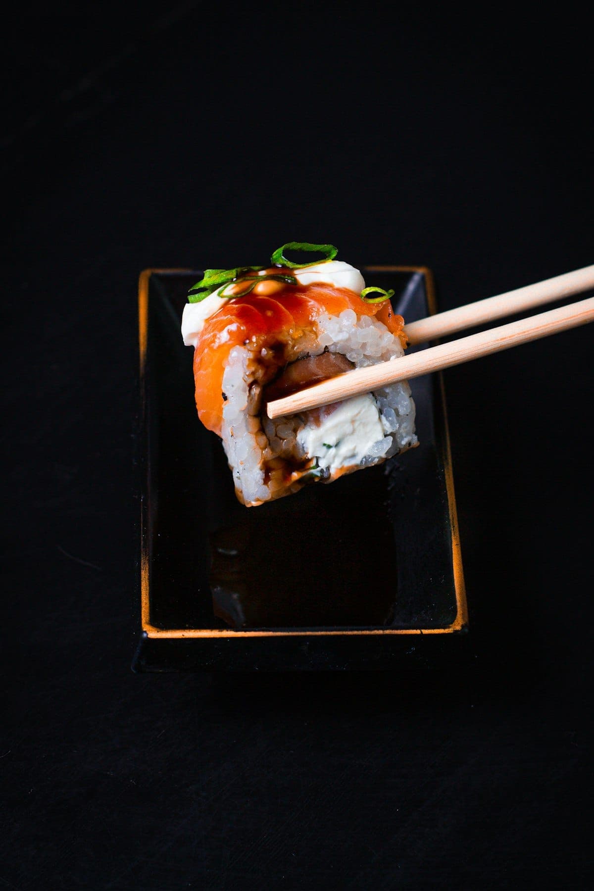 Maki saumon fromage saisi par des baguettes au-dessus d'une assiette noire laquée